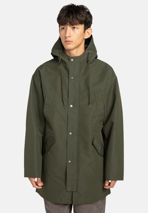 Jonge man in een donkergroen gewatteerd regenjack met drukknoopsluiting, klepzakken en trekkoorden, over grijze broek, kijkend naar de zijkant.