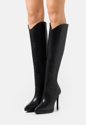 Schwarze kniehohe Lederstiefel mit spitzer Zehe, schlankem Design und hohem Stöckelabsatz. Am oberen Rand leicht eingesenkt.