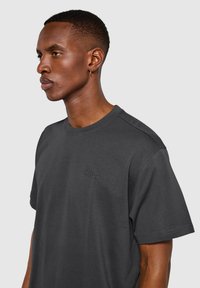 Mörkgrå t-shirt i bomull med rundad hals, korta ärmar och diskret präglad logotyp vid bröstet. Mjuk textur.