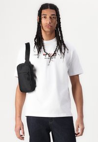Black shoulder bag made of fabric, worn over a white t-shirt with colorful text. Jeans feature a dark wash and visible stitching details.