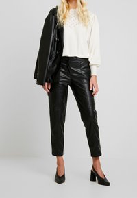 Maglione crema con accenti in perline testurizzate, abbinato a pantaloni di pelle neri aderenti. Il modello indossa anche un blazer nero e scarpe con tacco.
