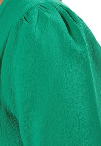 Groen textiel met een lichte glans, met een ontwerp van geplooide puffmouwen. Het oppervlak vertoont een gerimpeld patroon.