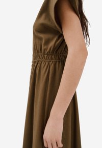 Abito verde oliva con maniche corte, fascia in vita arricciata e una texture di tessuto morbida. La silhouette presenta un design di gonna fluente.