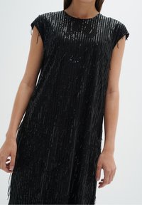 Černé šaty s krátkým rukávem, zdobené vertikálními flitry a třásněmi, vytvářejícími třpytivou texturu a vrstvený efekt.