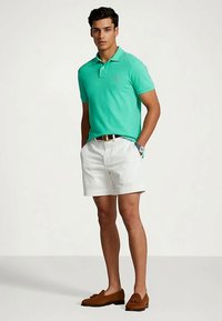 Man som bär en turkos polo tröja, vita shorts, brunt bälte och bruna loafers, stående med händerna i fickorna.