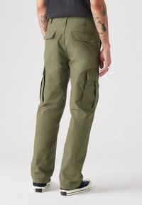 Calças cargo verde-oliva com bolsos de remendo e abas laterais, feitas de um material de algodão resistente. Ajustadas no tornozelo, combinadas com sapatilhas escuras.