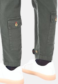 Pantaloni cargo verdi con tasche laterali, dettagli con bottoni e orli arrotolati, abbinati a sneaker bianche con suola rosa chiaro.
