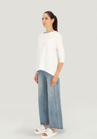 Biały, luźny top z długim rękawem połączony z szerokimi, jasnoniebieskimi dżinsami oraz białymi sandałami na płaskiej podeszwie, z okrągłym dekoltem i subtelnym detalem marki.