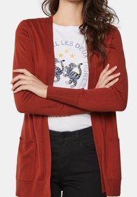 Kardigan v rezavé barvě s dlouhými rukávy, otevřeným předním dílem a dvěma předními kapsami, přes bílý tričko s potiskem a černé kalhoty.