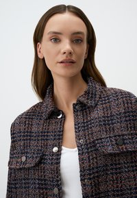 Veste en tweed avec une palette de couleurs bleu marine et orange, présentant un col, des boutons en métal argenté et deux poches avant.