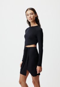 Crni uski top s dugim rukavima i izrezom, u kombinaciji s visokim omotnim suknjom. Glatka tkanina s elegantnim, minimalnim dizajnom.