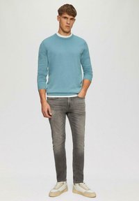 Hellblauer Rundhalspullover aus strukturiertem Stoff, getragen über einem weißen T-Shirt, kombiniert mit grauen Slim-Fit-Jeans und beigen Sneakers.