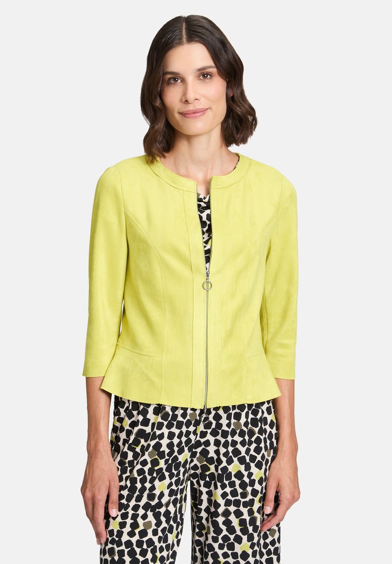 Jasnozielona kurtka z okrągłym dekoltem, rękawami o długości 3/4, zamkiem błyskawicznym z przodu i peplum na dole; zestawiona z czarno-białymi spodniami w wzór.