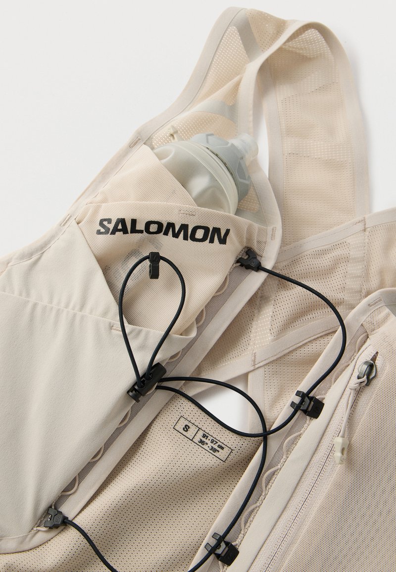 Gilet de course beige Salomon en tissu mesh, avec cordons noirs ajustables, poche zippée et une bouteille d'eau dans la poche avant. Étiquette taille petite visible.