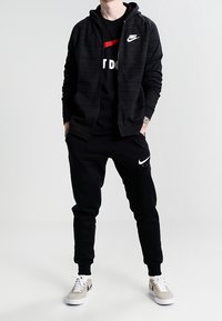 Svart Nike zip-up hoodie i texturerat material, kombinerad med en svart T-shirt med ett rött rand, och svarta joggers med vita detaljer.