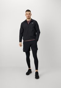 Veste légère noire avec une fermeture éclair à l'avant et un logo, associée à un short noir et un legging ajusté, portée avec des chaussures de sport noires.