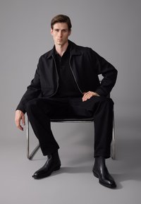 Chaqueta exterior negra con cierre frontal, llevada sobre una camiseta polo negra, combinada con pantalones negros y elegantes botas chelsea negras, sentado en una silla metálica.
