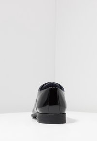 Chaussure formelle en cuir noir brillant vue de l'arrière, posée sur une surface blanche avec un fond gris clair uni.