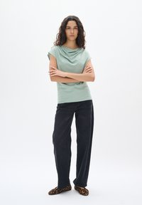 Top drappe mint verde con maniche corte, abbinato a pantaloni neri a gamba larga e pantofole con stampa leopardata, su sfondo bianco.