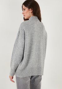 Harmaa neulepaita, jossa on korkea kaulus ja ribbetty helma. Suunnittelussa on oversized-leikkaus ja pehmeä rakenne.
