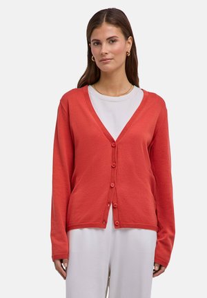 Donna che indossa un cardigan corallo con bottoni sopra una maglietta bianca e pantaloni bianchi, in piedi contro uno sfondo chiaro e uniforme.