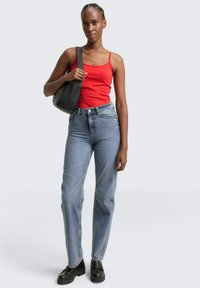 Rode mouwloze tanktop, hooggewaisted lichtblauwe jeans, zwarte handtas en zwarte platform schoenen. Eenvoudige achtergrond.