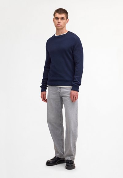 Ung mand iført en navyblå sweater, lysegrå jeans og sorte sko, stående mod en ensfarvet hvid baggrund.