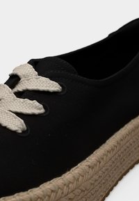 Μαύρο πάνινο αθλητικό παπούτσι με χονδρό πλεκτό σόλα από τζούτ και χονδρά εκρού κορδόνια δεμένα μέσα από μεταλλικά πατήματα.