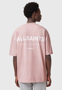 Camiseta de algodón rosa de gran tamaño con un llamativo texto blanco en la parte posterior, que incluye "ALLSAINTS" y "LONDRES" con caracteres japoneses debajo.