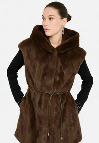Gilet in finta pelliccia marrone con cappuccio, caratterizzato da un motivo a onde testurizzato, vita con cintura, e una camicia nera a maniche lunghe con dettagli argento sui polsini.