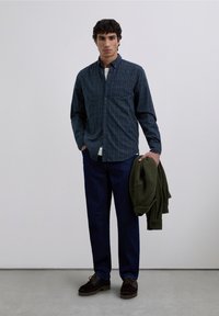 Camisa de cuadros azul y verde con botones, pantalones de mezclilla oscuros, sosteniendo un suéter verde, zapatos marrones, de pie contra un fondo neutro.