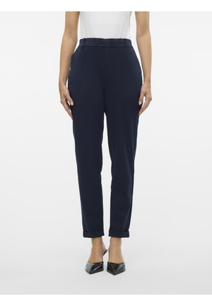 Vrouw die marineblauwe, hooggetailleerde, taps toelopende broek met omgeslagen zoom en zwarte pumps met puntige neus draagt, staand tegen een witte achtergrond.