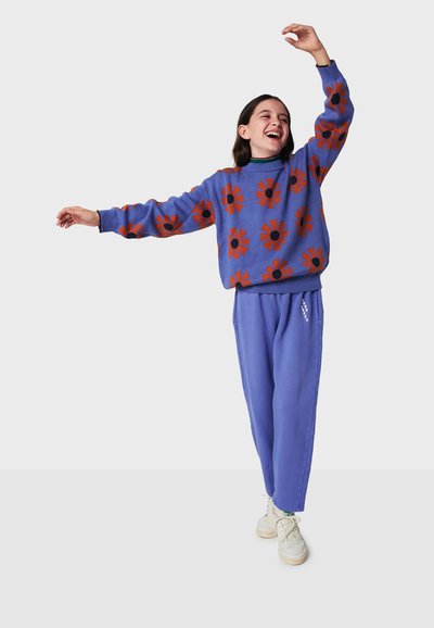 Maglione viola con motivo a fiori rossi, abbinato a pantaloni viola coordinati. La modella sorride e ha i capelli castani, indossando sneakers bianche.