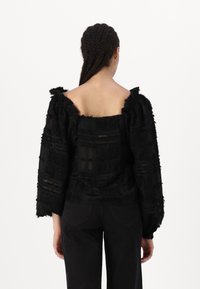 Schwarze strukturierte Bluse mit schulterfreier Gestaltung und Puffärmeln. Verfügt über schichtweisen Stoff und transparente Abschnitte. Getragen mit schwarzen Hosen.