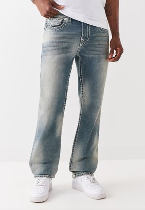 Mann, der ausgebleichte blaue Jeans und weiße Turnschuhe trägt, steht mit einer Hand auf der Hüfte und der anderen seitlich.