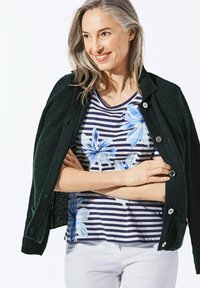 Schwarze strukturierte Strickjacke über einem gestreiften Hemd mit blauen Blumenmustern. Das Model hat graue Haare, verschränkte Arme und trägt weiße Hosen.