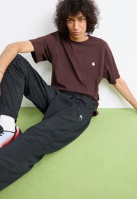Ung person med krøllet hår iført en brun t-shirt og sorte Nike-bukser, der sidder på et grønt tæppe op ad en hvid væg.