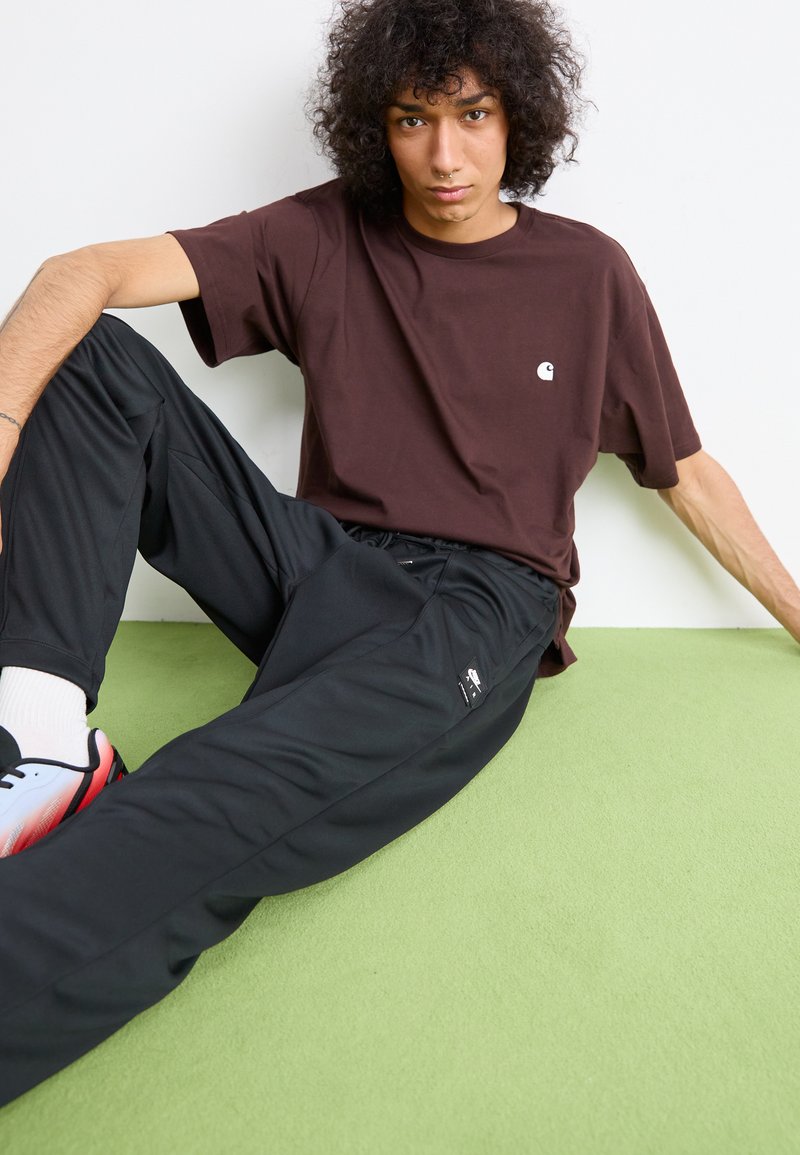 Giovane con capelli ricci che indossa una maglietta marrone e pantaloni neri Nike, seduto su un tappeto verde contro un muro bianco.