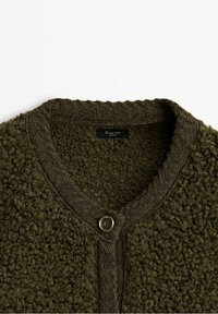 Gebreid vest in donkergroen. Voorzien van een gestructureerde boucle-stof met een gladde kraag en een goudkleurige knoopsluiting aan de voorkant.