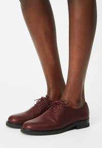 Jambes à la peau foncée portant des chaussures en cuir bordeaux à lacets avec semelles noires sur un fond blanc.