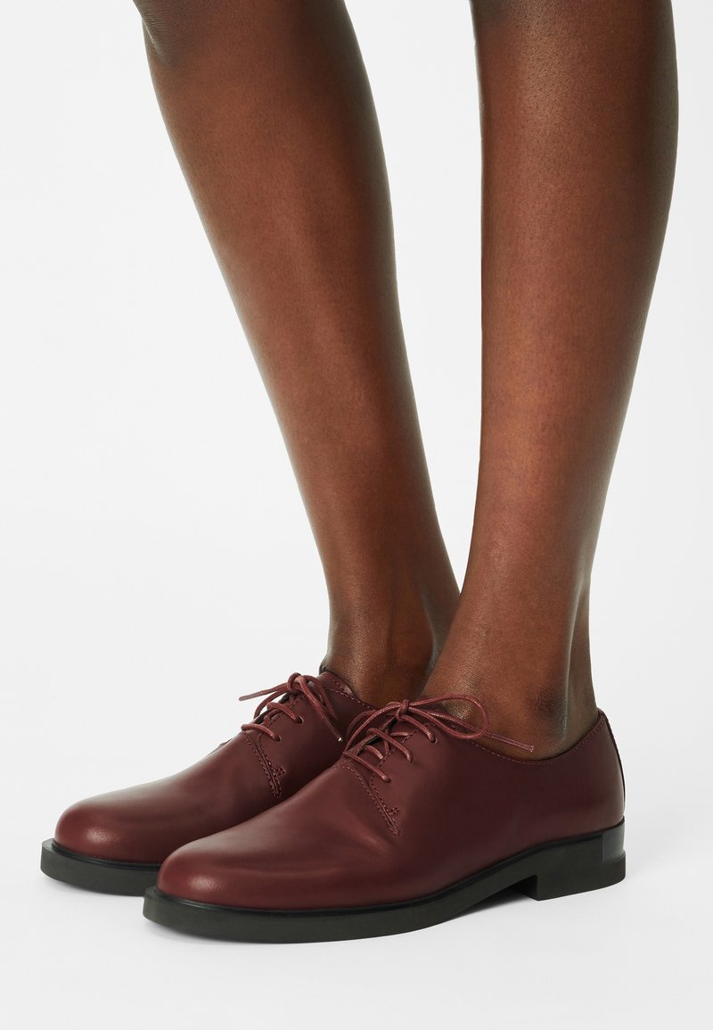 Jambes à la peau foncée portant des chaussures en cuir bordeaux à lacets avec semelles noires sur un fond blanc.