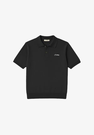 Młody mężczyzna w czarnym polo z białym napisem, szare spodnie w prążki, czarna czapka z daszkiem z pomarańczowym logo, trzymający brązowe rękawiczki w lewej ręce.