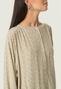 Blusa beige con pois bianchi, dettagli dei bottoni e tessuto morbido e leggero. Collo rotondo e maniche leggermente arricciate.