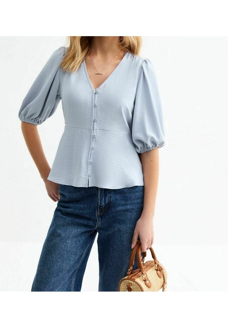 Femme portant un blouse à manches bouffantes bleu clair et un jean taille haute, tenant un petit sac à main en osier. Fond neutre.