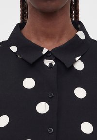 Κοντινό πλάνο μαύρου πουκάμισου με μεγάλα άσπρα πουά και μυτερό γιακά που φοράει ένα άτομο με πλεξούδες στα μαλλιά.