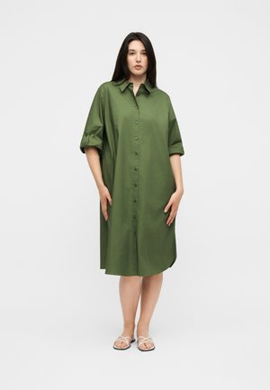 Femme aux longs cheveux foncés portant une robe midi ample boutonnée vert olive et des sandales blanches à brides, debout devant un fond clair uni.