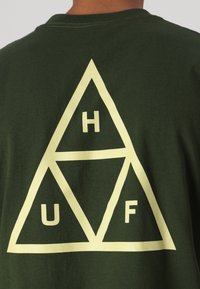 T-shirt en coton vert foncé avec un grand graphique triangulaire jaune, contenant les lettres H, U et F disposées dans différentes sections.