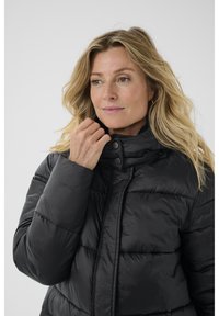 Chaqueta acolchada negra con cuello alto, cierre de cremalleras y acentos de botones a presión. Presenta una textura acolchada y un acabado ligero y brillante.