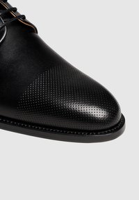 Chaussure en cuir noir avec une texture lisse et perforée, bout rond et coutures décoratives le long de la semelle. Design à lacets.