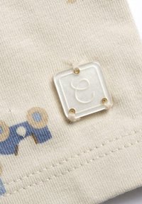 Tessuto di cotone beige con motivi circolari blu e marroni. Etichetta in plastica trasparente con la lettera 'e' in rilievo attaccata, con quattro fori visibili.